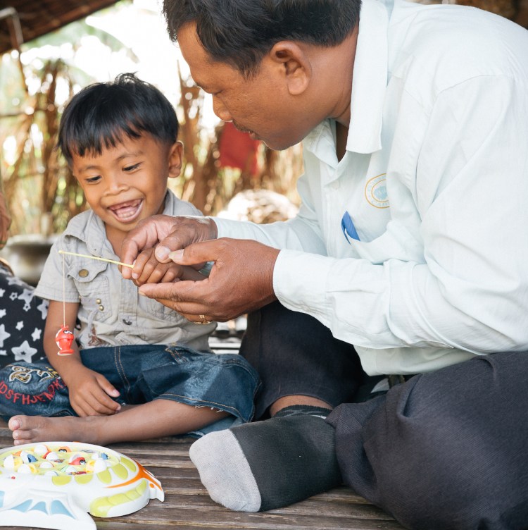 Four-year-old Makara shows a wide smile as he plays a game with Sokha, his disability worker. Makara has a communication disability, and Sokha is helping him overcome it. (Photo: Hugo Sharp/OIC: The Cambodia Project)